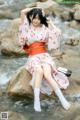 A woman in a pink kimono sitting on a rock by a river.