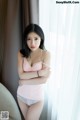 A woman in a pink tank top and panties posing by a window.
